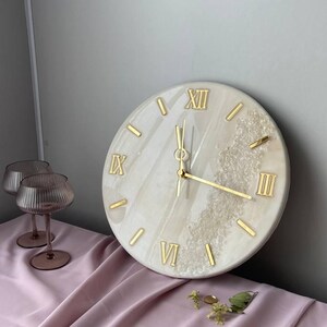Elegant handcrafted marble resin wall clock with Roman numerals and gold accents, placed on a soft pink fabric surface. Decorative glassware and a few sprigs of greenery complement the luxurious aesthetic of the clock.