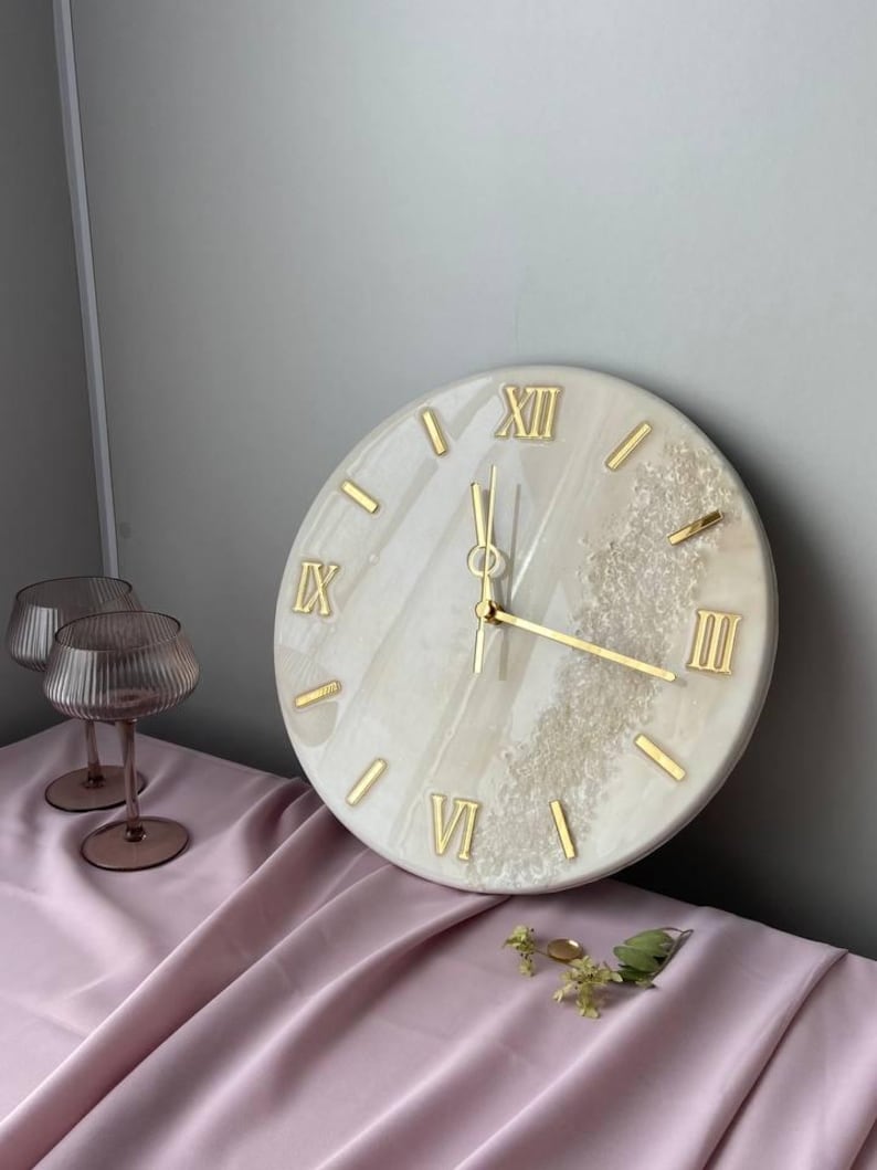 Elegant handcrafted marble resin wall clock with Roman numerals and gold accents, placed on a soft pink fabric surface. Decorative glassware and a few sprigs of greenery complement the luxurious aesthetic of the clock.