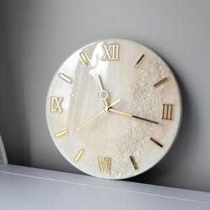 Puede incluir: Reloj de pared redondo con esfera blanca y números romanos dorados. Las manecillas y los números del reloj son dorados. El reloj presenta un diseño texturizado y cristalino en el lado derecho de la esfera.