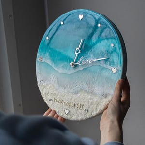 May include: A round resin clock featuring a blue and white ocean wave design. The clock face has silver hands and silver hearts. The clock is personalised with the name "YULIA OLEKSIY" and the word "love" in silver lettering.