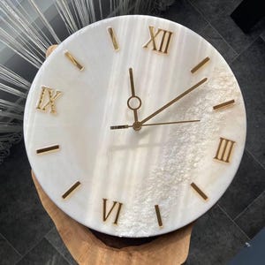 Elegant handcrafted marble resin wall clock with Roman numerals and gold accents, placed on a wooden surface.