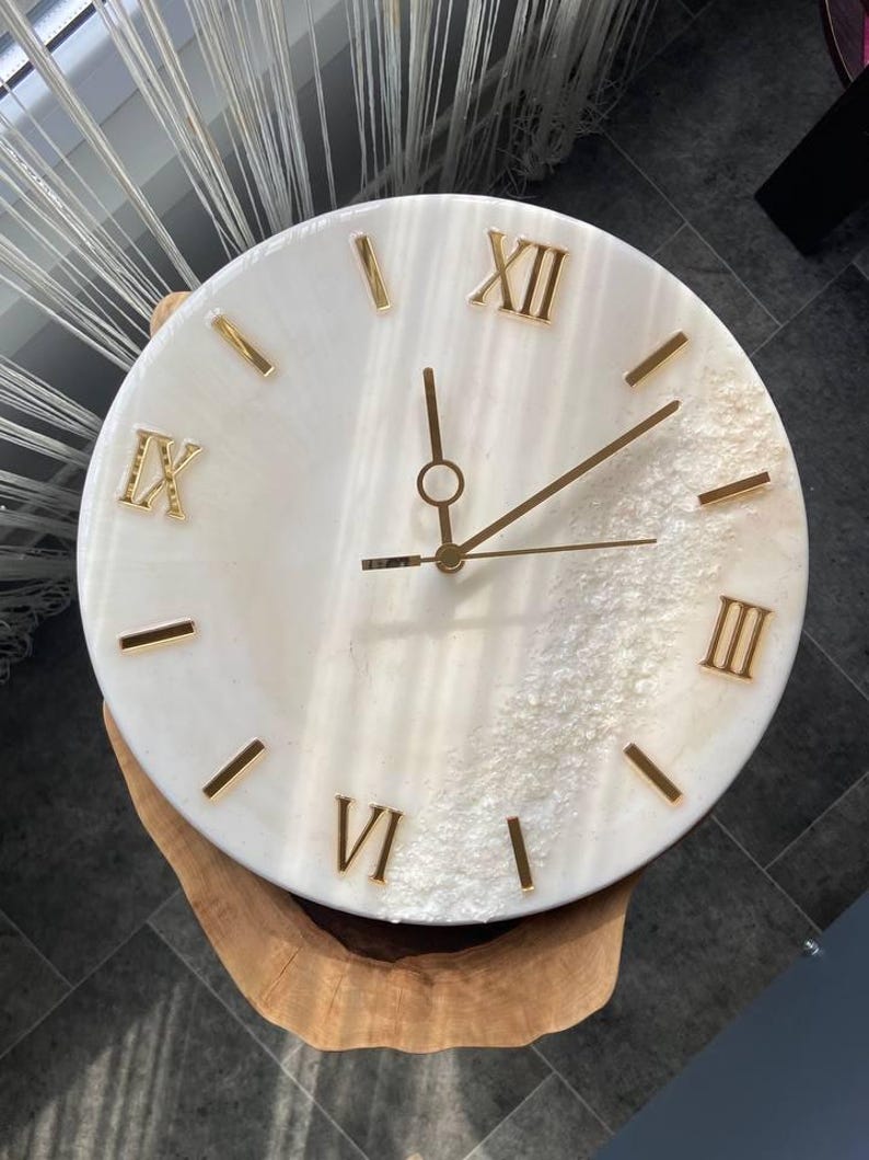 Elegant handcrafted marble resin wall clock with Roman numerals and gold accents, placed on a wooden surface.