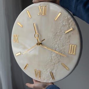 A person holding a handcrafted marble resin wall clock, showcasing Roman numerals and gold clock hands. The clock features a luxurious blend of ivory and grey tones with a textured resin finish.