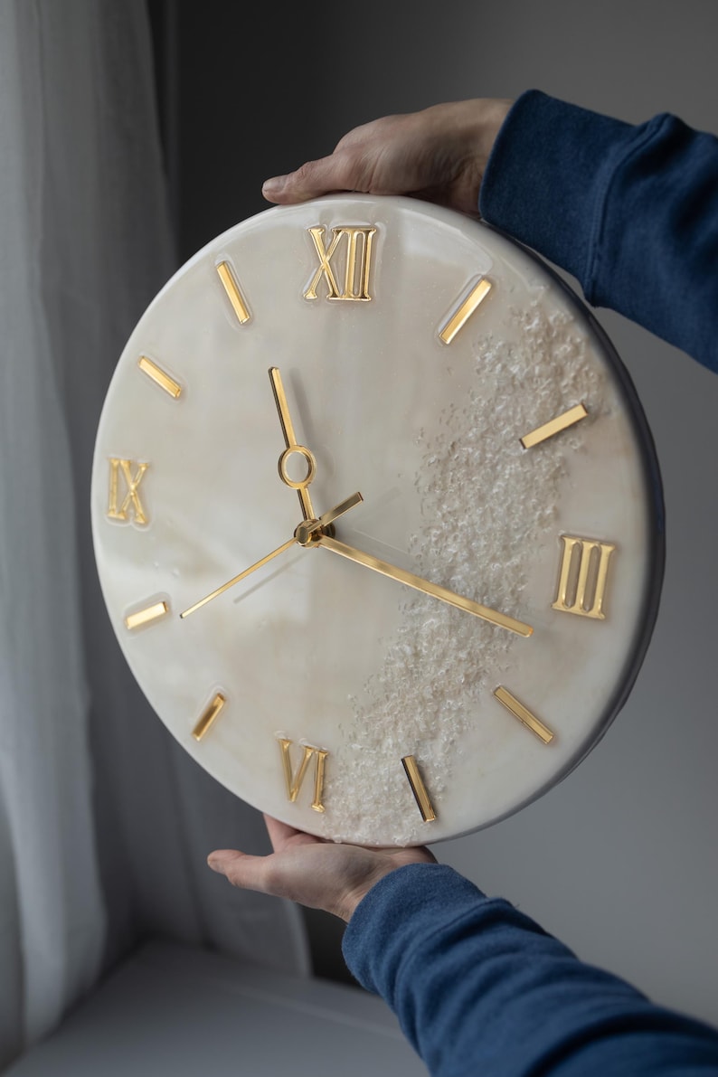 A person holding a handcrafted marble resin wall clock, showcasing Roman numerals and gold clock hands. The clock features a luxurious blend of ivory and grey tones with a textured resin finish.