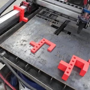 May include: Red plastic clamps used to hold a metal sheet in place on a metal cutting machine. The clamps are shaped like an L and have multiple holes for attaching to the sheet.
