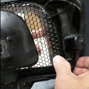 May include: Close-up of a black plastic car part with a honeycomb-shaped grille. A hand is holding the part, showing how it can be removed.