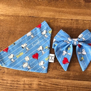 May include: Blue bandana with a yellow trim and a white Snoopy print. The bandana has the words "Smile", "Giggle", "Laugh", and "Fred & Juno" printed on it. A matching blue bow tie with a white Snoopy print and red hearts.