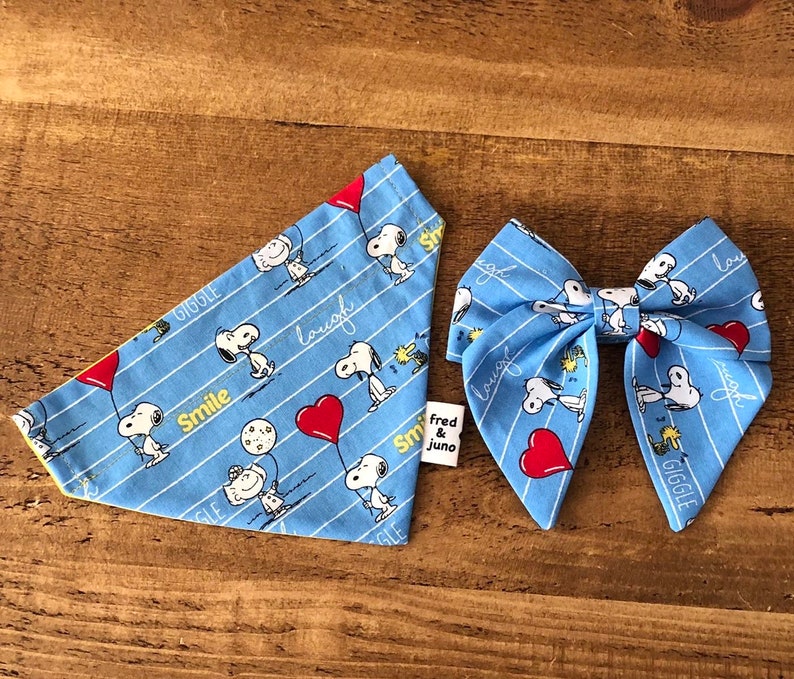 May include: Blue bandana with a yellow trim and a white Snoopy print. The bandana has the words "Smile", "Giggle", "Laugh", and "Fred & Juno" printed on it. A matching blue bow tie with a white Snoopy print and red hearts.