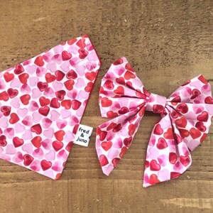 May include: A pink and red heart print dog bandana and bow tie set. The bandana has a white background with red and pink hearts. The bow tie is also white with red and pink hearts. The bandana has a label that says "fred & juno".