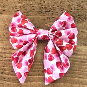 May include: A pink and red bow with a heart pattern. The bow is made of fabric and is tied in a knot.