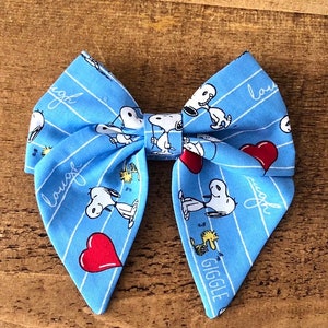 May include: A blue bow with a white stripe pattern featuring Snoopy and Woodstock. The bow has the words "laugh" and "giggle" printed on it.