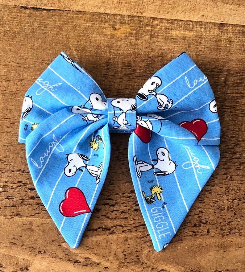 May include: A blue bow with a white stripe pattern featuring Snoopy and Woodstock. The bow has the words "laugh" and "giggle" printed on it.