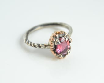 Puede incluir: Un anillo de plata con una banda retorcida y una piedra preciosa ovalada rosa. La piedra preciosa está engastada en un bisel dorado con un diseño texturizado. El anillo está sobre un fondo blanco.