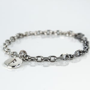May include: Silver-toned chain bracelet with a small, round charm. The charm features the letter "F" in a simple, sans-serif font. The bracelet is made of linked oval and round chain segments.