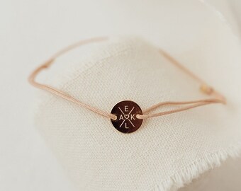 May include: A delicate bracelet with a small, round, gold-colored charm featuring the initials "E A K L". The bracelet is strung on a thin, light pink cord. The image is set against a white background with a cream-colored fabric and dried leaves.
