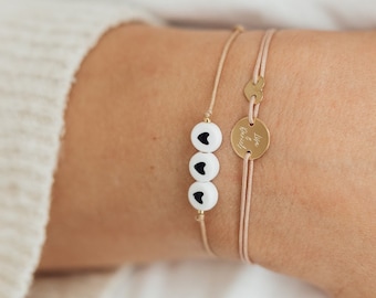 May include: Two delicate bracelets on a wrist. One features three white beads with black heart designs. The other has a small gold disc with an engraved initial. Both are strung on thin, light pink cords.