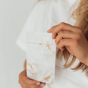 Puede incluir: Una persona sostiene una tarjeta blanca con un dise&ntilde;o floral y el texto "Du bist in meine Herzensschritte werden". Un charm de coraz&oacute;n dorado est&aacute; unido a una cuerda fina.