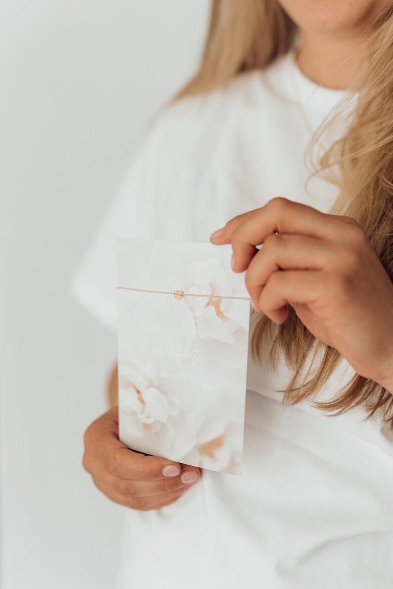 Puede incluir: Una persona sostiene una tarjeta blanca con un dise&ntilde;o floral y el texto "Du bist in meine Herzensschritte werden". Un charm de coraz&oacute;n dorado est&aacute; unido a una cuerda fina.