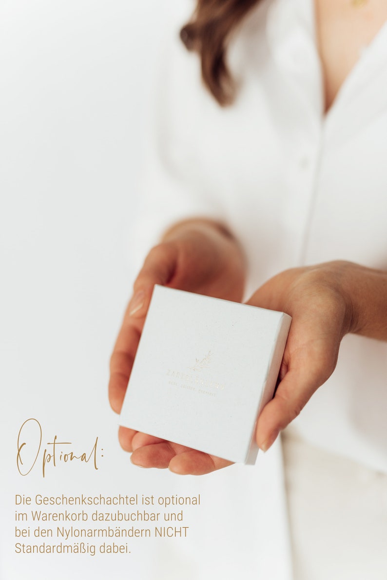 Puede incluir: Una caja de regalo blanca con un peque&ntilde;o dise&ntilde;o de hoja y el texto "Zarte Sch&auml;tze" en letras doradas. El texto "Optional: Die Geschenkschachtel ist optional im Warenkorb dazubuchbar und bei den Nylonarmb&auml;ndern NICHT Standardm&auml;&szlig;ig dabei." est&aacute; escrito debajo de la caja.