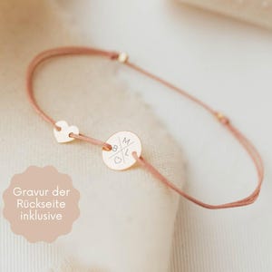 Könnte beinhalten: Ein zartes Armband mit einer dünnen, staubrosa farbenen Kordel. Es hat einen kleinen Herzanhänger und einen runden, goldfarbenen Anhänger mit eingravierten Initialen. Der Text auf dem Bild lautet "Gravur der Rückseite inklusive".