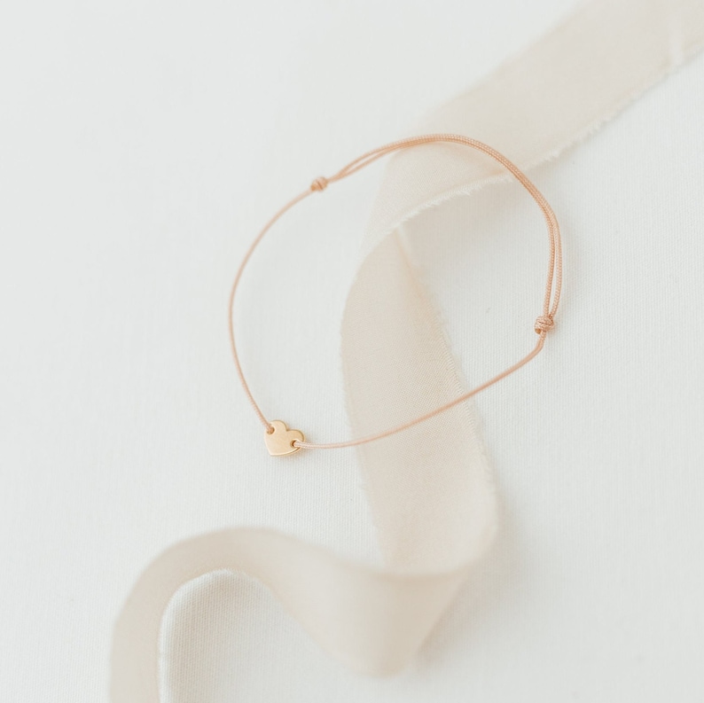 Puede incluir: Un delicado brazalete de coraz&oacute;n de oro con un cord&oacute;n fino de color marr&oacute;n. El brazalete se muestra sobre un fondo blanco con una cinta beige.