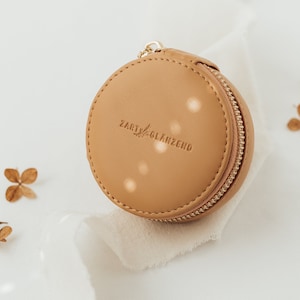 May include: A round, tan leather zippered pouch with the text "ZART GLANZEND" embossed on the front. The pouch has a gold-colored zipper and a small loop. Dried flowers and a white fabric are in the background.