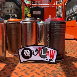 以下が含まれることがあります： 黒い円の中にスタイリッシュな文字Qと上向きの矢印が入った、黒と赤の文字で「OUTLAW AL DIAZ」と書かれた白いパッチ。パッチはスプレー缶の列の前にある。