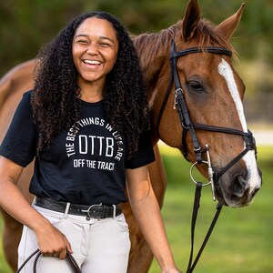 May include: A person wearing a black t-shirt with the text "THE BEST THINGS IN LIFE ARE OTTB OFF THE TRACK" stands next to a brown horse with a white blaze. The person is smiling and holding a riding crop.