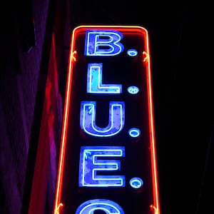 Puede incluir: Un letrero de neón con borde rojo y letras de neón azules que deletrean "B.L.U.E.S."