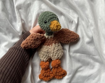 Puede incluir: Un juguete de pato hecho a mano con ganchillo, con cabeza verde, alas marrones y patas naranjas. El pato tiene un pico amarillo y naranja y un pecho color crema. El juguete está hecho de hilo suave y texturizado.