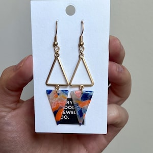 May include: A pair of gold triangle earrings with a blue, orange, and white abstract pattern. The earrings are hanging from a white card with the text "Very Cool Jewel Co."