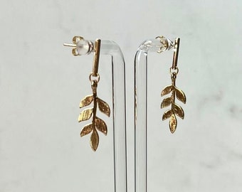 May include: Gold-colored leaf earrings displayed on a clear acrylic stand. The earrings have a delicate, detailed design, resembling small branches with multiple leaves. The background is a light marble pattern.