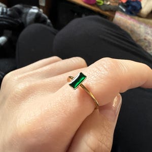 May include: A gold ring featuring a rectangular, emerald-green gemstone. The ring's band is thin and appears to be gold-colored. The gemstone is set in a gold bezel, creating a minimalist and elegant design.
