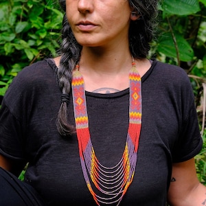 Könnte beinhalten: Eine farbenfrohe Perlenkette mit geometrischem Design. Die Halskette hat rote, orangefarbene, gelbe und violette Perlen, mit Strängen aus silbernen Perlen, die von unten herabhängen. Die Halskette wird vor einem schwarzen Oberteil getragen, mit einem grünen Hintergrund.