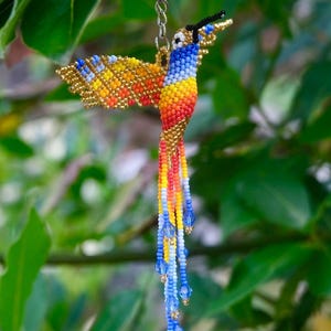 May include: A hummingbird keychain crafted from small, multi-colored beads in shades of yellow, orange, red, white, and blue. The bird features a long, slender beak, outstretched wings, and a tail composed of long, beaded strands with blue crystals.