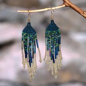 May include: A pair of handmade beaded earrings. The earrings have a dark teal beaded top with a fringe of green, blue, and gold beads. The earrings are suspended from a small branch.
