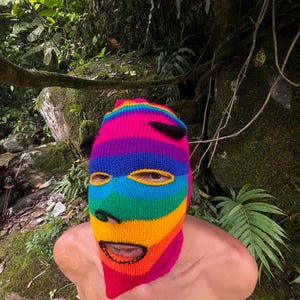 Puede incluir: Un pasamontañas de punto con rayas horizontales en colores vibrantes del arcoíris, incluyendo rosa, morado, azul, verde, amarillo y naranja. La prenda incluye dos pequeñas protuberancias negras en forma de cuerno en la parte superior y aberturas para los ojos y la boca.