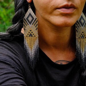 May include: Long beaded earrings with a geometric design. The earrings feature a diamond pattern in black and gold, with a fringe of gold, silver, and black beads. The earrings are hanging from a gold hook.