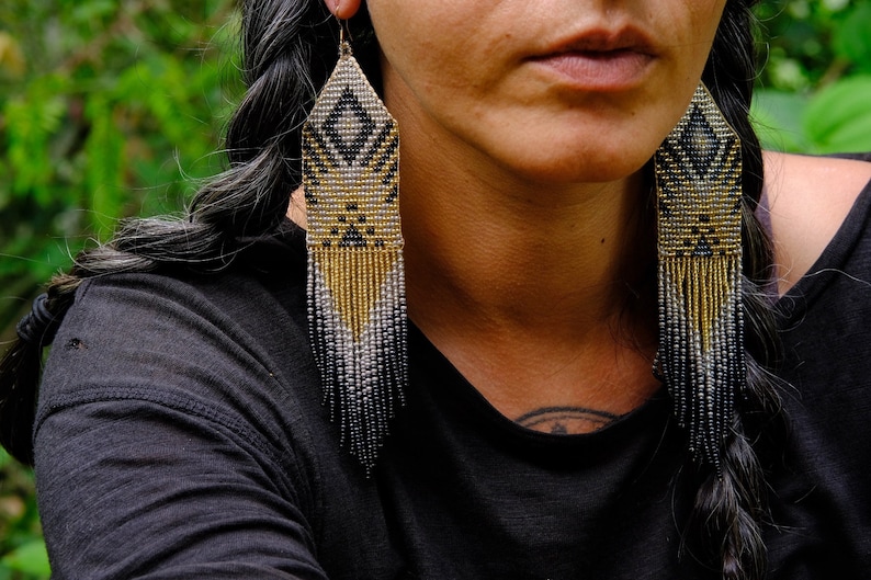 May include: Long beaded earrings with a geometric design. The earrings feature a diamond pattern in black and gold, with a fringe of gold, silver, and black beads. The earrings are hanging from a gold hook.