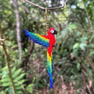 Pode incluir: Um chaveiro de arara de contas com uma cauda e asas coloridas do arco-íris. A arara é vermelha com um bico preto e um olho branco.