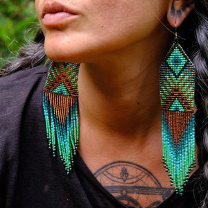 May include: Long, beaded earrings in shades of teal, green, and copper. The earrings have a geometric design at the top and a fringe of beads that hangs down. The earrings are worn by a person with dark hair.