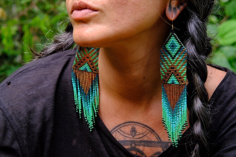 May include: Long, beaded earrings in shades of teal, green, and copper. The earrings have a geometric design at the top and a fringe of beads that hangs down. The earrings are worn by a person with dark hair.