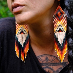 May include: Handmade beaded earrings with a geometric design. The earrings feature a pattern of orange, yellow, white, and black beads, with long fringe in shades of orange, red, and teal. The earrings are a statement piece.