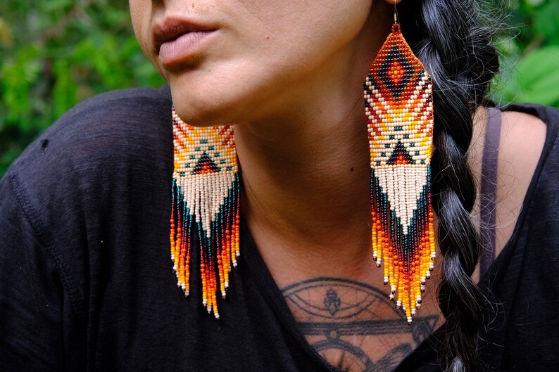 May include: Handmade beaded earrings with a geometric design. The earrings feature a pattern of orange, yellow, white, and black beads, with long fringe in shades of orange, red, and teal. The earrings are a statement piece.