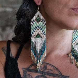May include: Handmade beaded earrings with a geometric design. The earrings feature a rectangular top with a pattern of green, white, and black beads. Long fringe in white, teal, and gold beads hangs from the bottom, creating a striking visual effect.