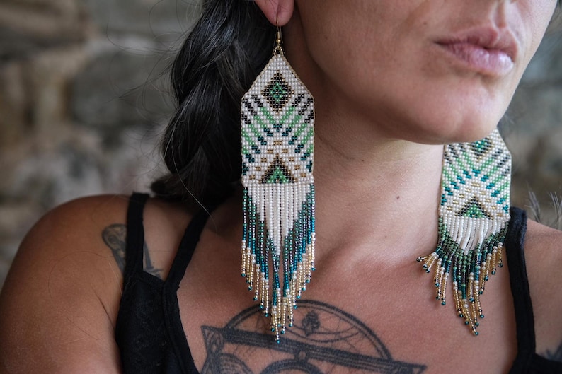 May include: Handmade beaded earrings with a geometric design. The earrings feature a rectangular top with a pattern of green, white, and black beads. Long fringe in white, teal, and gold beads hangs from the bottom, creating a striking visual effect.
