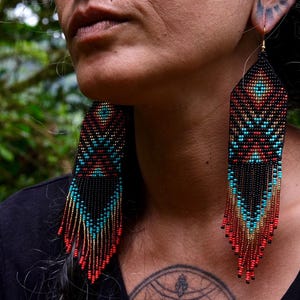 May include: Close-up of beaded earrings with a geometric design. The earrings feature a black base with turquoise, red, and gold accents. The fringe hangs down, creating a striking visual effect. The earrings are a unique accessory.