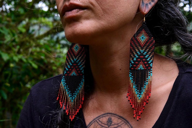 May include: Close-up of beaded earrings with a geometric design. The earrings feature a black base with turquoise, red, and gold accents. The fringe hangs down, creating a striking visual effect. The earrings are a unique accessory.