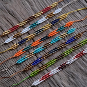 Op de afbeelding: Een verzameling kralenarmbanden in verschillende kleuren, waaronder rood, wit, goud, blauw, oranje en groen. De armbanden zijn diagonaal geplaatst op een verweerd houten oppervlak, wat hun ingewikkelde patronen en vakmanschap laat zien.