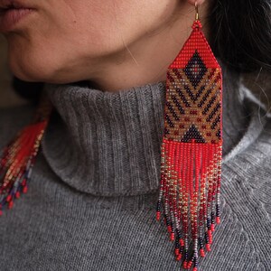 May include: Long, beaded earrings with a geometric design. The earrings feature a red, brown, and black pattern with dangling fringe. The earrings are worn with a gray turtleneck sweater.
